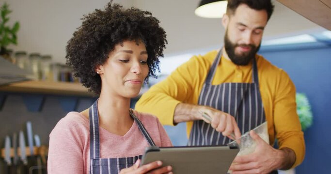 Video of happy diverse couple in aprons using tablet and baking in kitchen at home