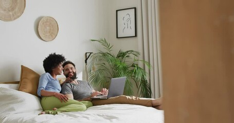 Video of happy diverse couple relaxing at home, lying on bed using laptop and smartphone, copy space - Powered by Adobe