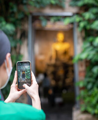 Female hand use smartphone for taking photo with The Gold Buddha statue in a church covered with 100-year-old banyan tree at Wat Bangkung.
