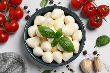 Delicious mozzarella balls in bowl, tomatoes and basil leaves on white marble table, flat lay