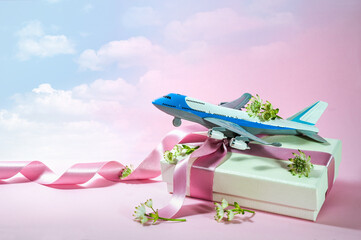 Toy airplane on a gift box with ribbon and some flowers, pink background fading into a cloudy blue sky, travel and journey present for Valentines day or wedding trip, copy space - 560292132