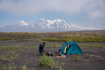 Coast of Pacific Ocean. Man and woman live in tent on a beach with black sand. There three volcanoes in the background.