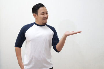 Young Asian man wearing white shirt smiling, showing and pointing something on his side, against white wall with copy space