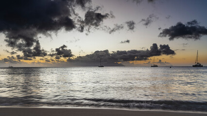 Evening seascape. Picturesque clouds in the sky, highlighted in orange. Silhouettes of yachts and islands on the horizon. Waves are foaming on a sandy beach. Long exposure. Seychelles. Mahe. 