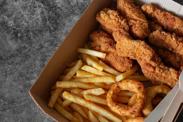 Breaded fried chicken breast with fries and onion rings, in a cardboard tray with wooden cutlery....