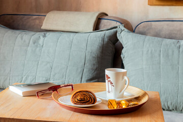 On the coffee table in the living room, a cup of morning tea with a fresh poppy seed bun. Women's reading glasses, a book.