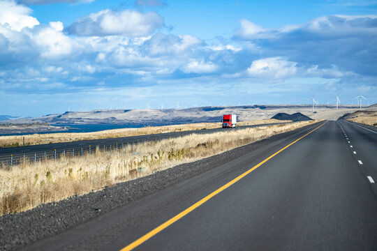 Landscaping with red big rig semi truck with dry van semi trailer driving on the divided highway road with electric wind turbines on the background
