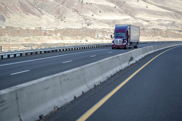 Purple big rig semi truck with high cab transporting cargo in dry van semi trailer running on the wide highway road with bare hills on the background