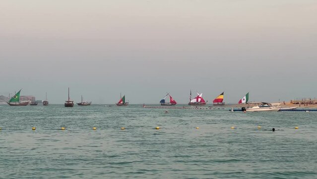boats with world cup countries' flag on it pass.Katara beach.some people swim and some watching. sunset time. weather become cool and tourists leave the beach. Doha, Qatar 2022.