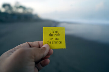 Business motivational and life inspirational quote - Take the risk or lose the chance. With person holding yellow card with motivation text in hand on beach background.