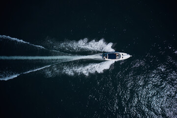 A white motor boat with a blue awning moves quickly on dark water, top view.