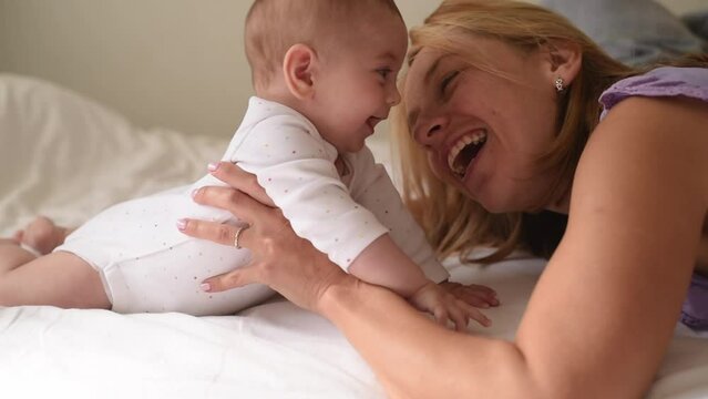 Smiling baby and mother lies on belly on white bed. Woman kiss and laugh with baby. Love and care in happy family. Side view. 