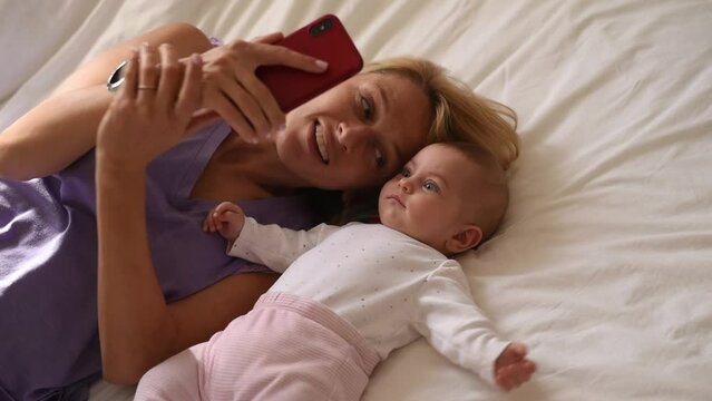 Mother and baby lies on white bed. Woman holds phone in hand, talks by video call and kiss child. Kid looks with interest and extends hand. Happy family, girls make selfie together at home.
