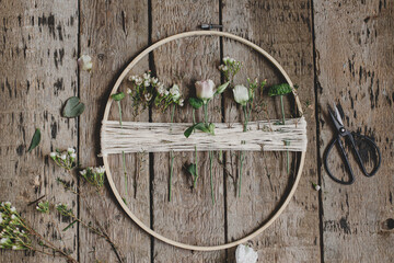 Making stylish spring wreath with beautiful fresh flowers. Wooden hoop, thread, scissors, and flowers on rustic wooden background flat lay. Modern and creative floral handmade decor
