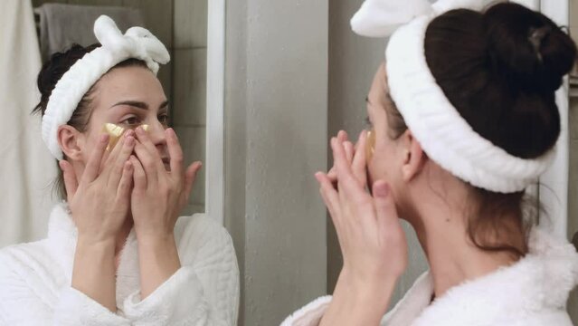 Close-up of a beautiful young woman with smooth healthy skin applying golden skin care patches under her eyes in the bathroom. cosmetic concept, slow motion