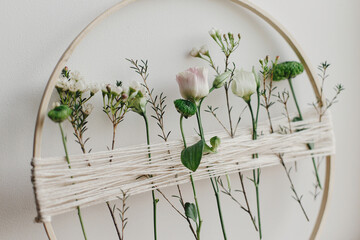 Stylish spring boho wreath with beautiful fresh flowers. Wooden hoop with flowers and thread on white wall background. Modern and creative floral handmade decor
