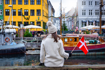 A tourist woman holding a Danish flag enjoys the view of the beautiful Nyhavn area in Copenhagen, Denmark, during winter time