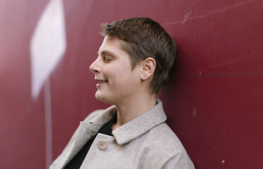 close-up portrait of young caucasian smiling lady with closed eyes and short hair lying against a red wall outside - female people with androgynous looking