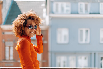 portrait beautiful smiling curly hair Brazilian young lady wearing sunglasses and orange color shirt looking in camera - outside home terrace picture in sunny daylight
