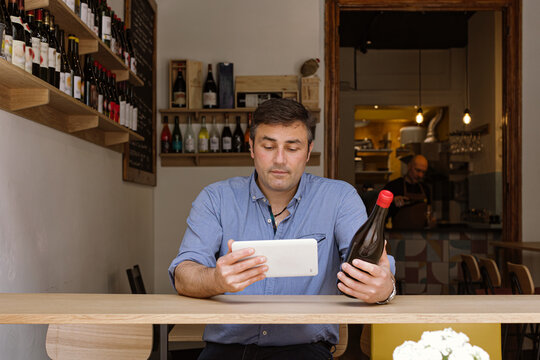 manager sitting in his wine shop and wine bar checks the label of a bottle of wine using his tablet - Powered by Adobe