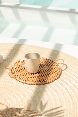 The mug is placed on a rattan tray. Swimming pool area. Decoration. Summer time. Vacation. Beach house. Relaxing space. Scandinavian style. Holiday vacation. Sunlight. Cozy space.