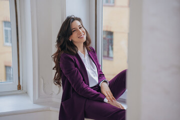 Gorgeous brunette  hispanic woman in violet suit sits on windowsill smiling wide looks at camera, satisfied female manager having fun, completed all tasks. Successful businesswoman with wavy hair home