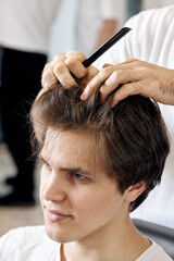 close-up, professional male hairstylist combing young customer's hair in barbershop