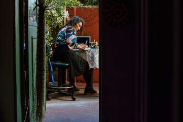 Fototapeta na wymiar Concentrated female artist working on a drawing on her terrace, seen from inside the house.