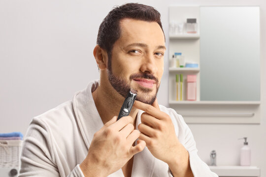 Young man in a bathrobe using a beard trimmer in a bathroom
