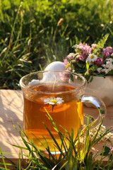 Cup of aromatic herbal tea, pestle and ceramic mortar with different wildflowers on green grass outdoors