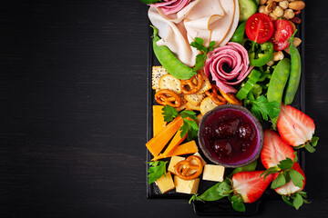 Fototapeta na wymiar Antipasto platter cold sliced ham, salami, crackers, strawberries, vegetables and cheese platter on board over dark background. Appetizers table with italian antipasti snacks. Top view