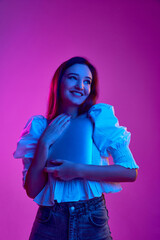 Portrait of young smiling girl, student in white blouse posing with laptop over purple background in neon light. Concept of emotions, youth, lifestyle