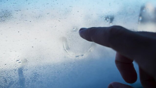 Close up on hand drawing a smilie face on glass window with condensation. Cold winter time as person draws a smiling face on wet glass