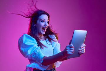 Portrait of young emotive girl cheerfully looking on tablet over pink background in neon light. Concept of emotions, youth, lifestyle - obrazy, fototapety, plakaty