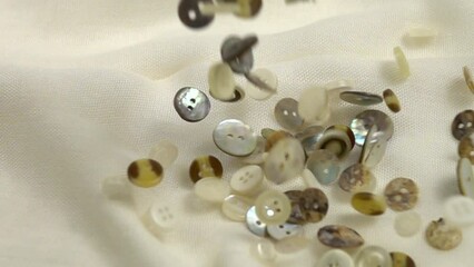 Small mother-of-pearl buttons for sewing to clothes are falling on white fabric, slow motion
