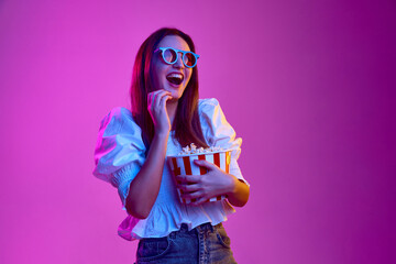 Comedy, laugh. Portrait of young emotive girl posing in 3D glasses with popcorn basket over pink...