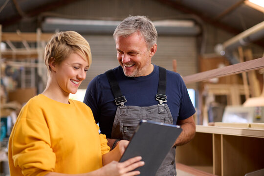 Female Apprentice With Digital Tablet Learning From Mature Male Carpenter In Furniture Workshop