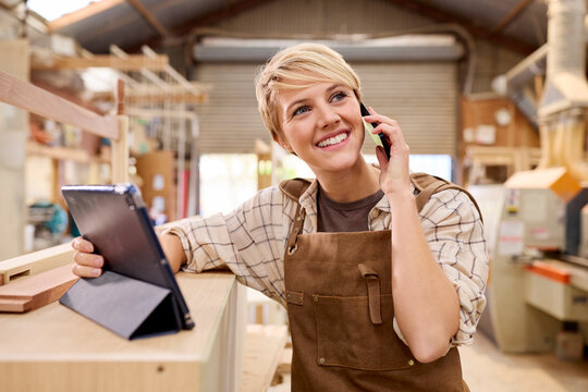 Female Apprentice With Digital Tablet Working As Carpenter In Furniture Workshop Making Phone Call