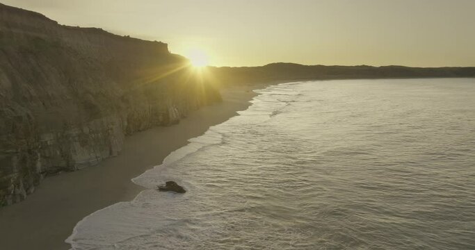 Sunrise peaks behind Fortrose cliffs with waves crashing on beach, aerial