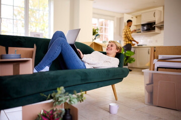 Couple Unpacking Boxes After Moving Into New Home Together As Woman Lies On Sofa Using Laptop