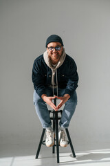 Model in hat and glasses is sitting on a chair. Handsome man is in the studio against white background