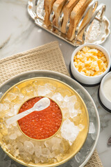 Orange Keta Lachs Kaviar in Dose auf Eis in Glas Schale mit Perlmutt Löffel, gekochte Eier Würfel, Charlotten Zwiebel Würfel, Toast Brot und Saure Sahne auf Serviette und Marmor hell