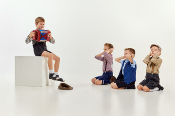 Fototapeta na wymiar Boy, child sitting and playing little accordion to his friends covering ears. Children over grey studio background. Concept of game, childhood, friendship