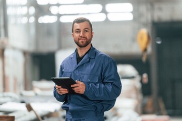 With tablet in hands. Factory worker in blue uniform is indoors - obrazy, fototapety, plakaty