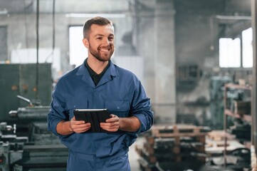 With tablet in hands. Factory worker in blue uniform is indoors - Powered by Adobe