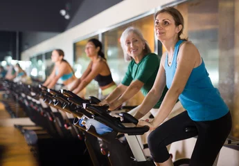 Foto op Plexiglas Fitness Positive mature female riding exercise bike during cycling class in modern gym