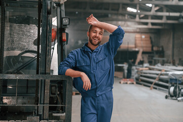 Leaning on the forklift. Factory worker in blue uniform is indoors