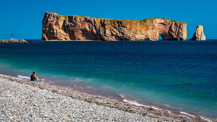 Woman sitting alone on a beach with the Perce Rock in the background.