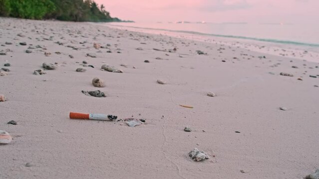 Burning fuming smoldering cigarette butt falling on sand at quite empty uninhabited beach at dawn pollution garbage closeup slow motion