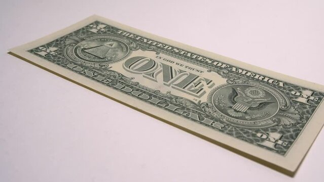 One new one dollar bill lies on a white surface with the reverse side. The camera rotates around the banknote.